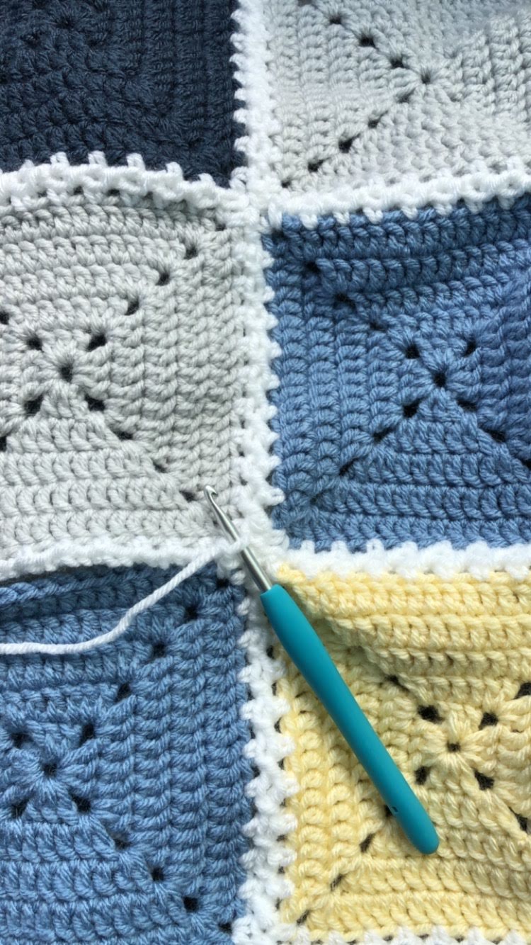 A crocheted blanket made up of blue and white squares arranged in a checkerboard pattern with a crochet hook laying on top.