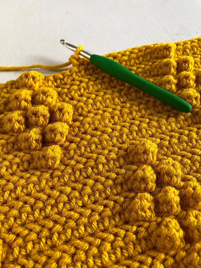 A crochet hook with a green base laying on top of a gold-colored crocheted blanket